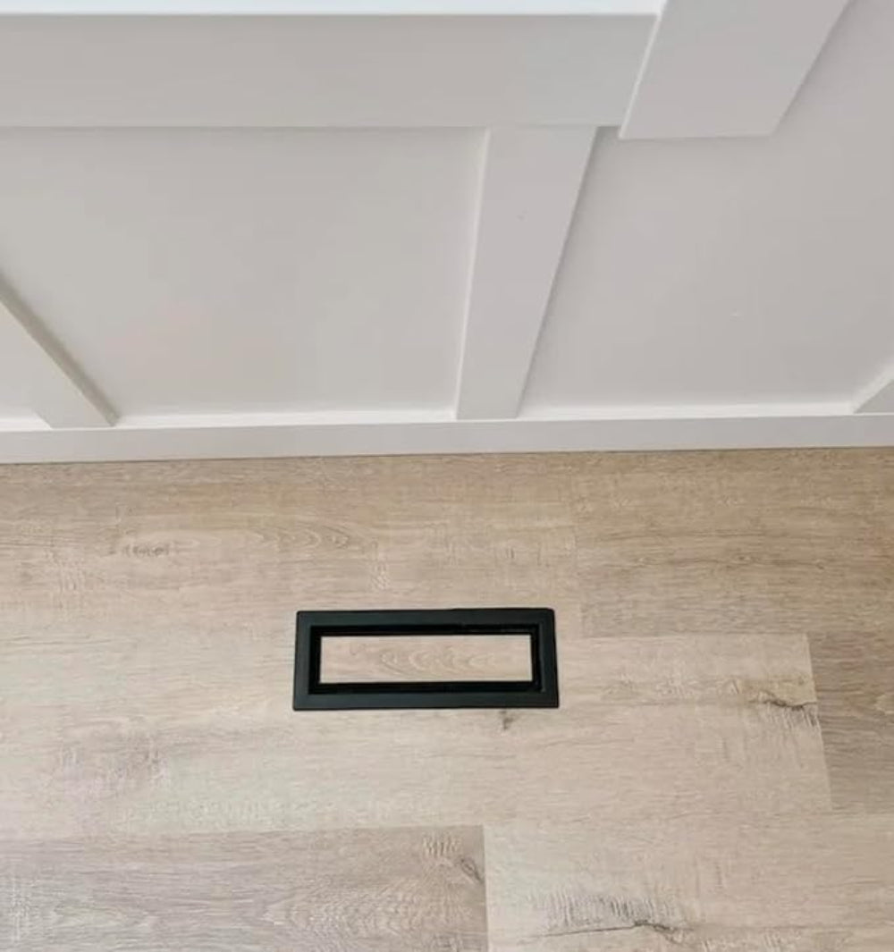 Black drop-in metal floor vent installed flush in hardwood flooring against a white baseboard — Khan Supply CO